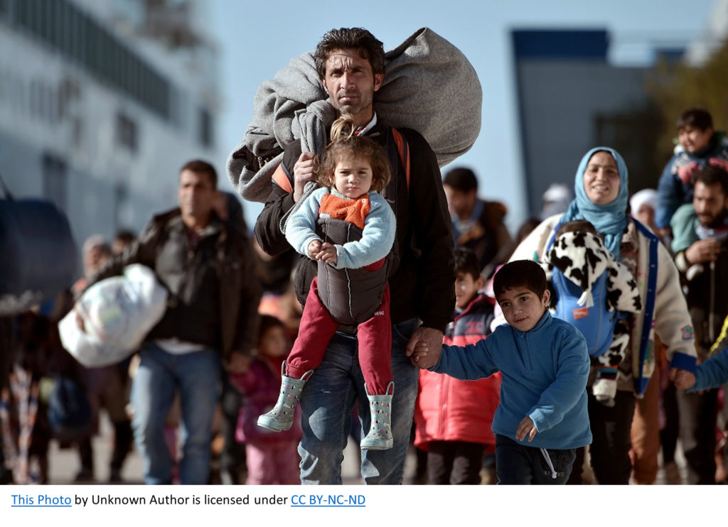 An Afghan man holds a young child in a child carrier followed by a number of other refugees.